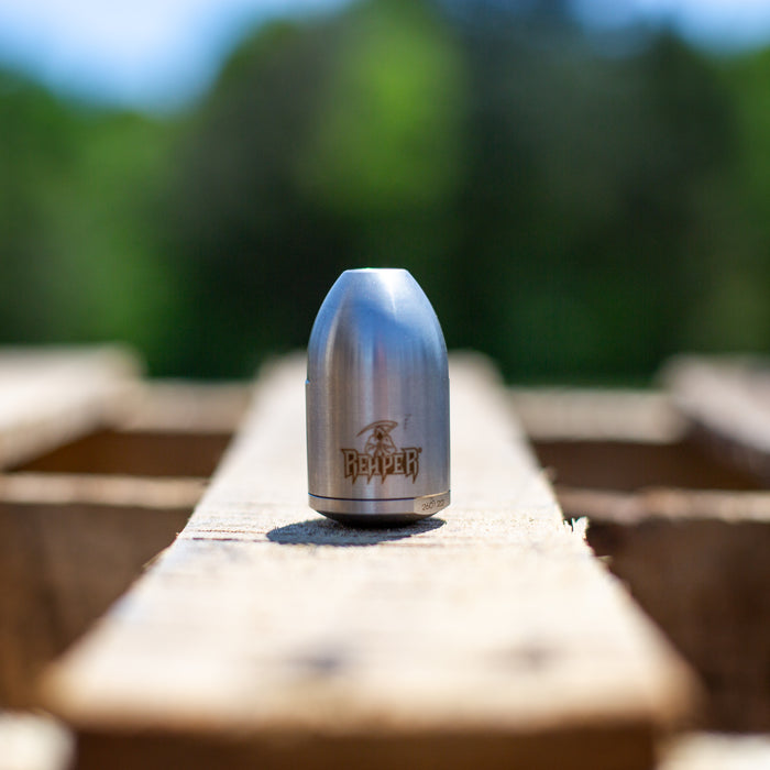Polished stainless steel 1/4-inch Reaper rotating sewer jetting nozzle standing upright on a wooden surface outdoors, engraved with Reaper logo and model marking. High‑pressure drain cleaning nozzle for 2–4 inch pipes featuring rotating front cutting jet and four rear propulsion jets, built with durable carbide wear surfaces for harsh environments.