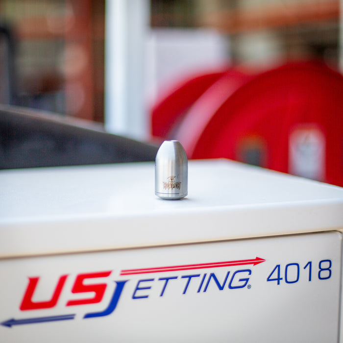 Stainless steel 1/4-inch Reaper rotating sewer jetting nozzle resting on a US Jetting 4018 machine, showing engraved Reaper logo and polished heavy‑duty construction. High‑pressure drain cleaning nozzle for 2–4 inch pipes with rotating front cutting jet and four rear propulsion jets, built with durable carbide wear surfaces for harsh environments.
