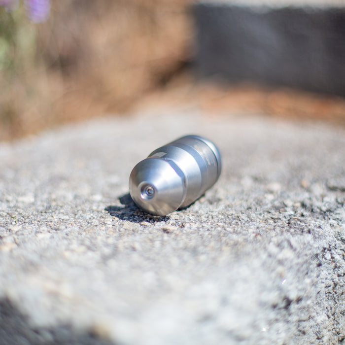 Compact stainless steel ENZ Bulldog‑style sewer jetting nozzle on a stone surface outdoors, featuring a precision‑machined rounded head with a central jet orifice and an oil‑free braking system for controlled slow rotation; compatible with fresh or recycled water and engineered for cutting grease, roots, and heavy pipeline deposits using powerful rear and forward jet patterns, with an optional 90‑degree descaling head available for removing tough scale from pipe walls.