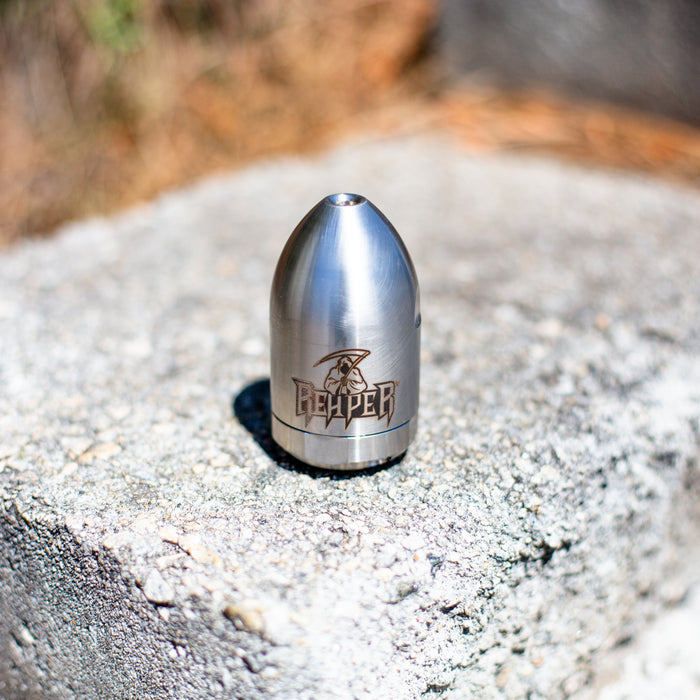 Reaper Rotating Nozzle placed on an outdoor wooden surface, featuring a polished stainless‑steel body with the engraved Reaper logo. This high‑pressure sewer jetting nozzle uses a powerful forward oscillating jet and four rear propulsion jets to cut blockages, penetrate roots, and clear grease with a cone‑shaped spray pattern. Precision‑machined design ideal for professional drain cleaning, available in multiple pressure and flow sizes from US Jetting.