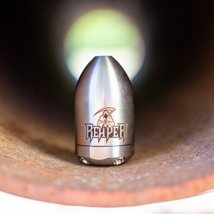 Reaper Rotating Nozzle positioned inside a rusted pipe, featuring a polished stainless‑steel body with the engraved Reaper logo. This high‑pressure sewer jetting nozzle delivers a powerful forward oscillating jet and four rear propulsion jets to cut blockages, penetrate roots, and clear grease with a cone‑shaped spray pattern. Precision‑machined design ideal for professional drain cleaning, available in multiple pressure and flow sizes from US Jetting.