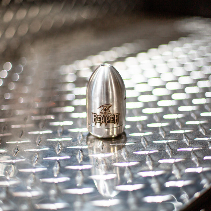 Reaper Rotating Nozzle displayed on a diamond‑plate surface, featuring a polished stainless‑steel body with the engraved Reaper logo. This high‑pressure sewer jetting nozzle uses a powerful forward oscillating jet and four rear propulsion jets to cut blockages, penetrate roots, and clear grease with a cone‑shaped spray pattern. Ideal for professional drain cleaning and available in multiple pressure and flow sizes from US Jetting.