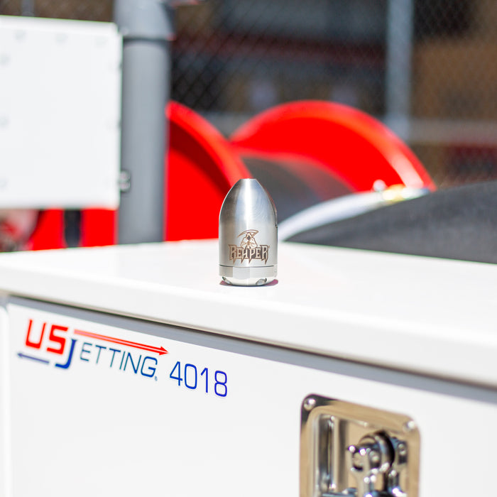 Reaper Rotating Nozzle resting on a US Jetting 4018 jetter, showing its stainless‑steel body with the REAPER logo and precision jet ports. This high‑pressure sewer jetting nozzle features a powerful forward oscillating jet and four rear propulsion jets designed to cut blockages, penetrate roots, and clear grease with a cone‑shaped spray pattern. Ideal for professional drain cleaning and available in multiple pressure and flow sizes from US Jetting.