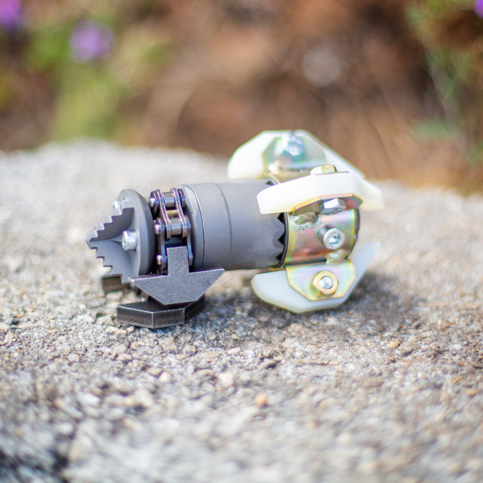 Close‑up of an ENZ Turbo Chain Scraper nozzle on a concrete surface, featuring a heavy‑duty toothed cutting head, rotating chain arm, adjustable chain links, and white centralizing skids. Industrial sewer jetting tool engineered for removing concrete, roots, and grease from 3–8 inch pipes, built for rugged conditions and compatible with jetting machines requiring at least 8 GPM.